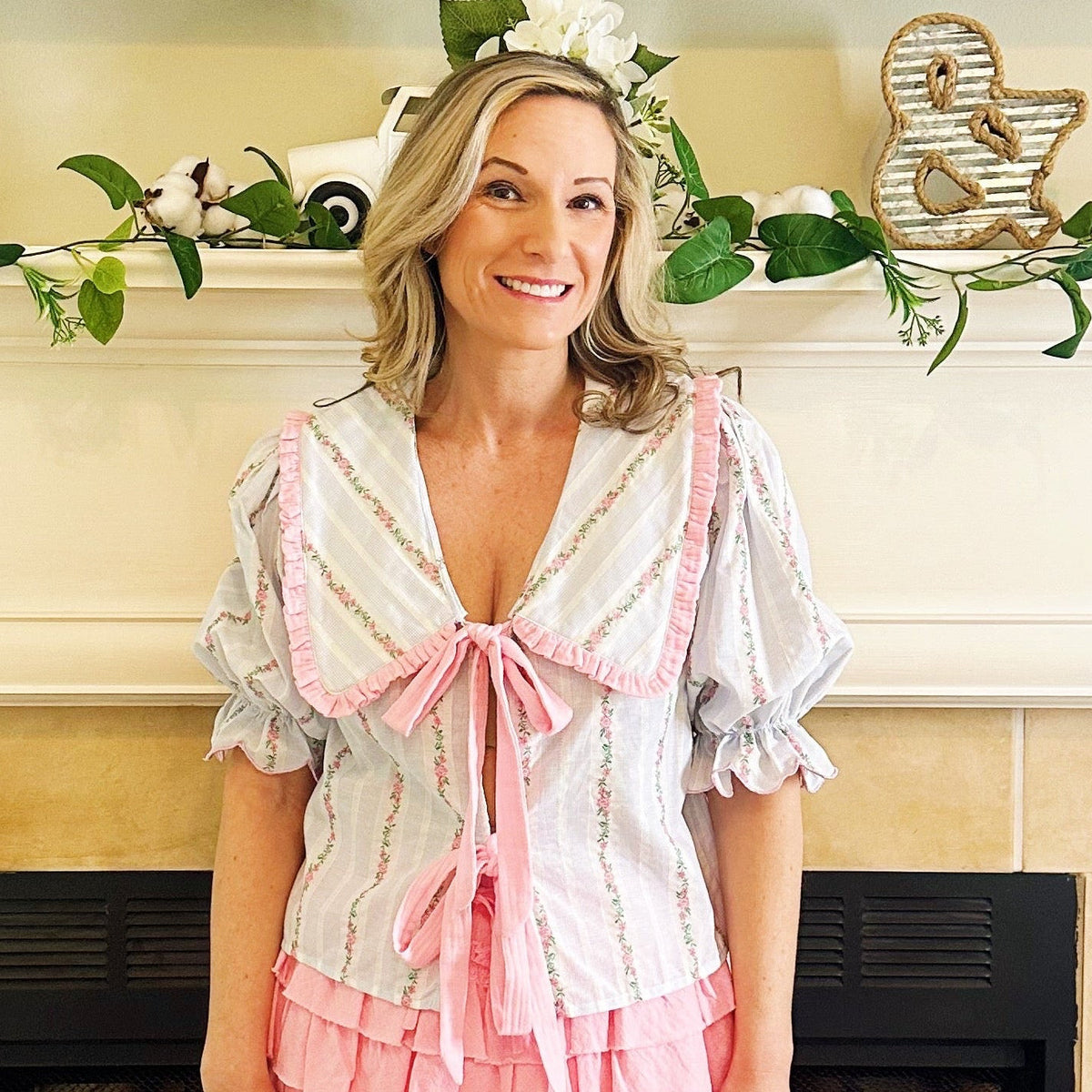 A woman stands in front of a fireplace wearing a short white blouse with pink ruffle trim and a pink tiered mini skirt, paired with white western boots. The fireplace mantel has greenery and decorative items, and the floor is tiled.