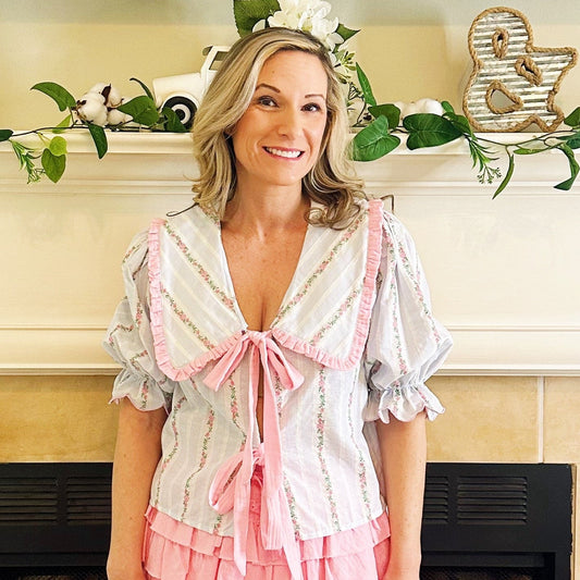 A woman stands in front of a fireplace wearing a short white blouse with pink ruffle trim and a pink tiered mini skirt, paired with white western boots. The fireplace mantel has greenery and decorative items, and the floor is tiled.