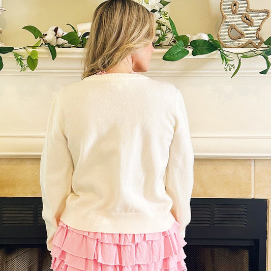 A woman stands with her back to the camera in front of a fireplace, wearing a cream knit sweater, a pink ruffled tiered mini skirt, and white cowboy boots.