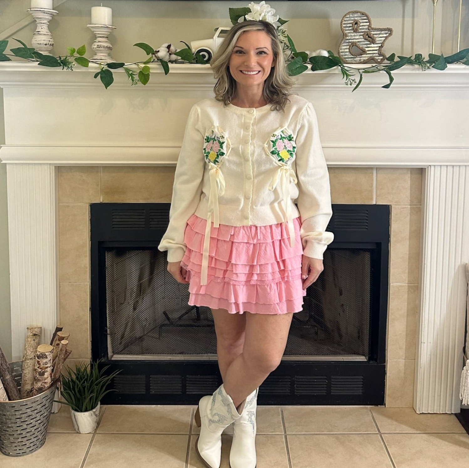 Woman in a white blouse with floral designs and a pink skirt standing in front of a fireplace.
