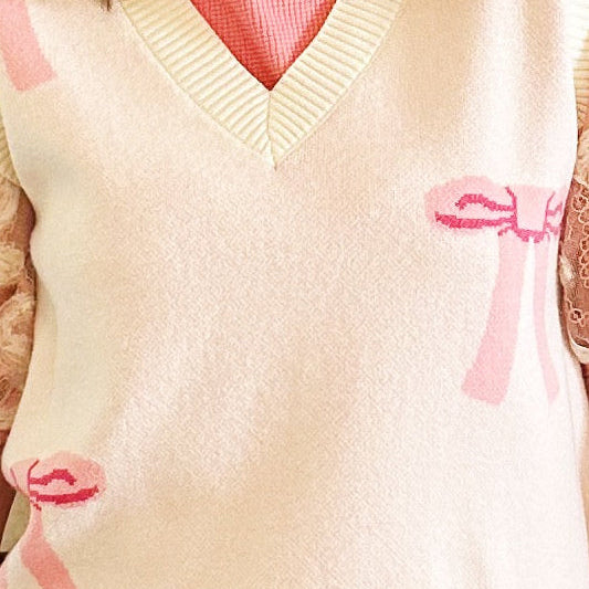 A woman wearing a cream knit sweater vest with pink bow details, sheer lace puff sleeves, and a pink skirt, standing in front of a fireplace.