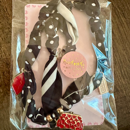 A handmade pet collar set in a clear plastic bag, featuring a black and white polka dot collar, a black and white striped collar, a black plaid collar, and red enamel purse charms on a pink backing card.