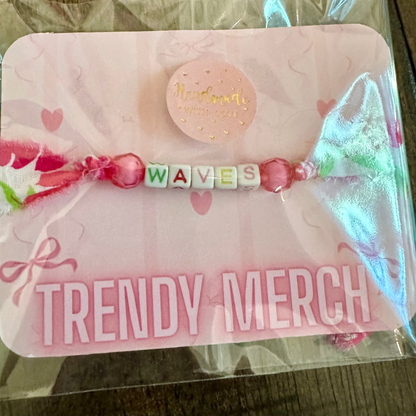 A pink beaded bracelet spelling 'WAVES' with pink beads and a central round bead, displayed on a pink backing card with 'TRENDY MERCH' printed on it, inside a clear plastic sleeve.