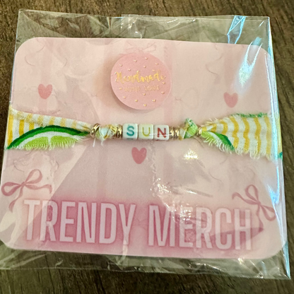 A handmade beaded bracelet with a yellow and white striped ribbon bow, displayed on a pink backing card labeled 'TRENDY MERCH' inside a clear plastic sleeve, set on a dark wood background.
