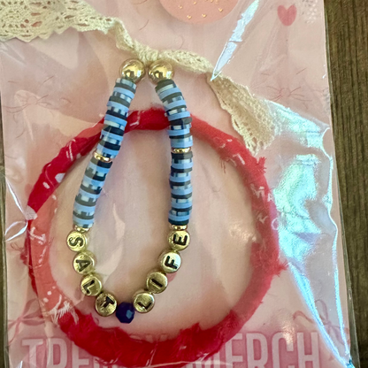A beaded bracelet set displayed on a pink retail card with lace trim and a round logo sticker. The set includes a red elastic bracelet, a blue-and-white striped elastic bracelet, and gold-tone beaded accents spelling out names, all wrapped in clear plastic.