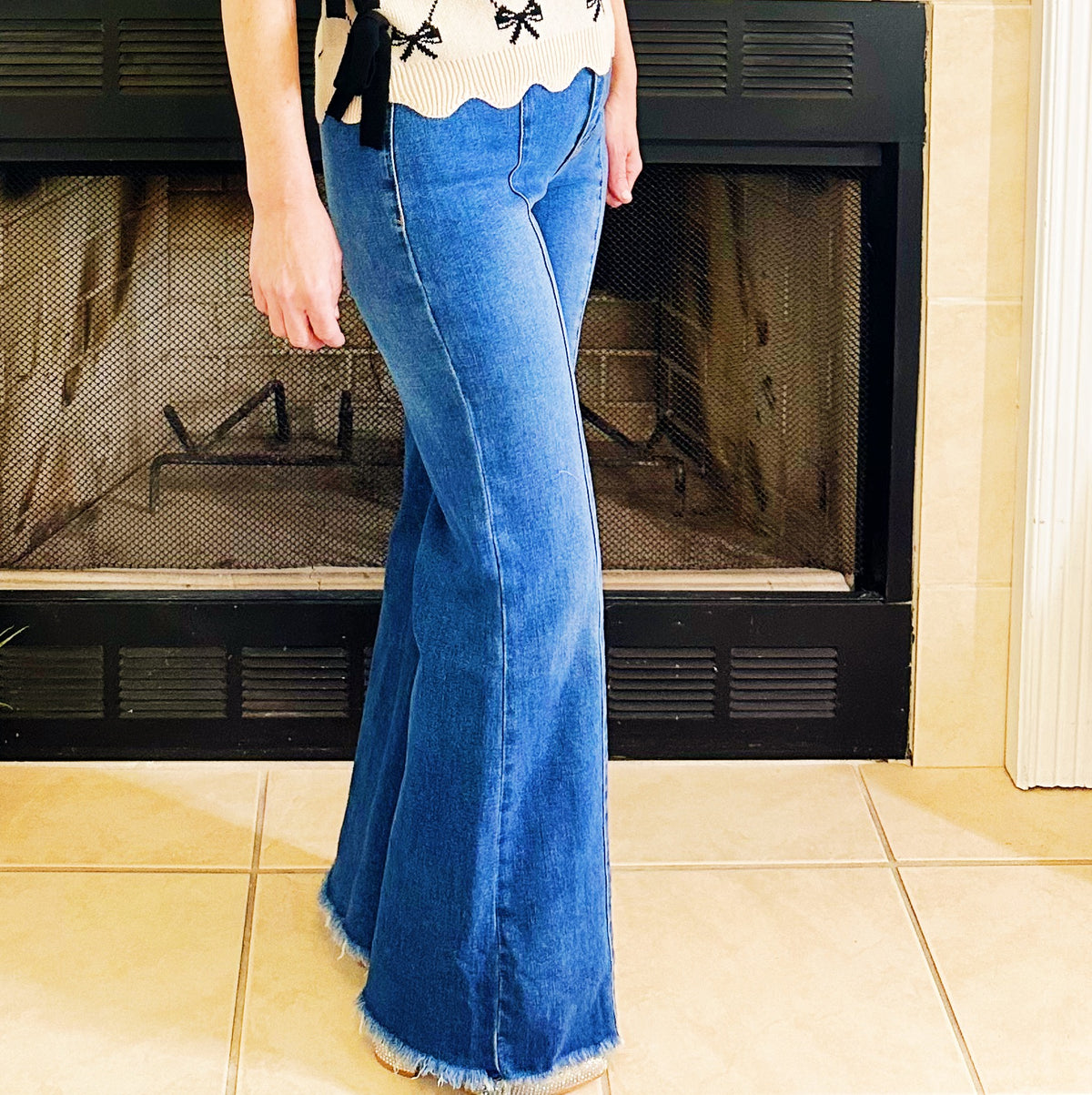 A person wearing a pair of medium-wash blue flare jeans with a frayed hem, standing in front of a black fireplace with beige tile flooring.
