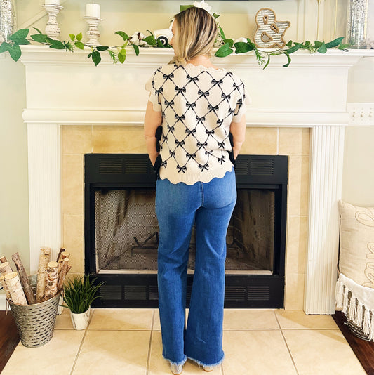 A woman stands with her back to the camera in front of a fireplace, wearing a cream short-sleeve top with a black bow pattern and scalloped hem, paired with blue jeans.
