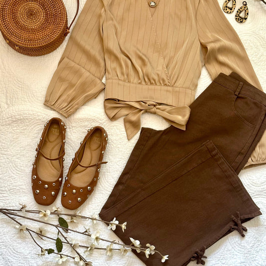 A styled flat-lay outfit set featuring a beige long-sleeve ribbed blouse with a tie waist, dark brown pants with decorative ties at the hem, brown studded flats, and a round woven straw bag, arranged on a white textured blanket.
