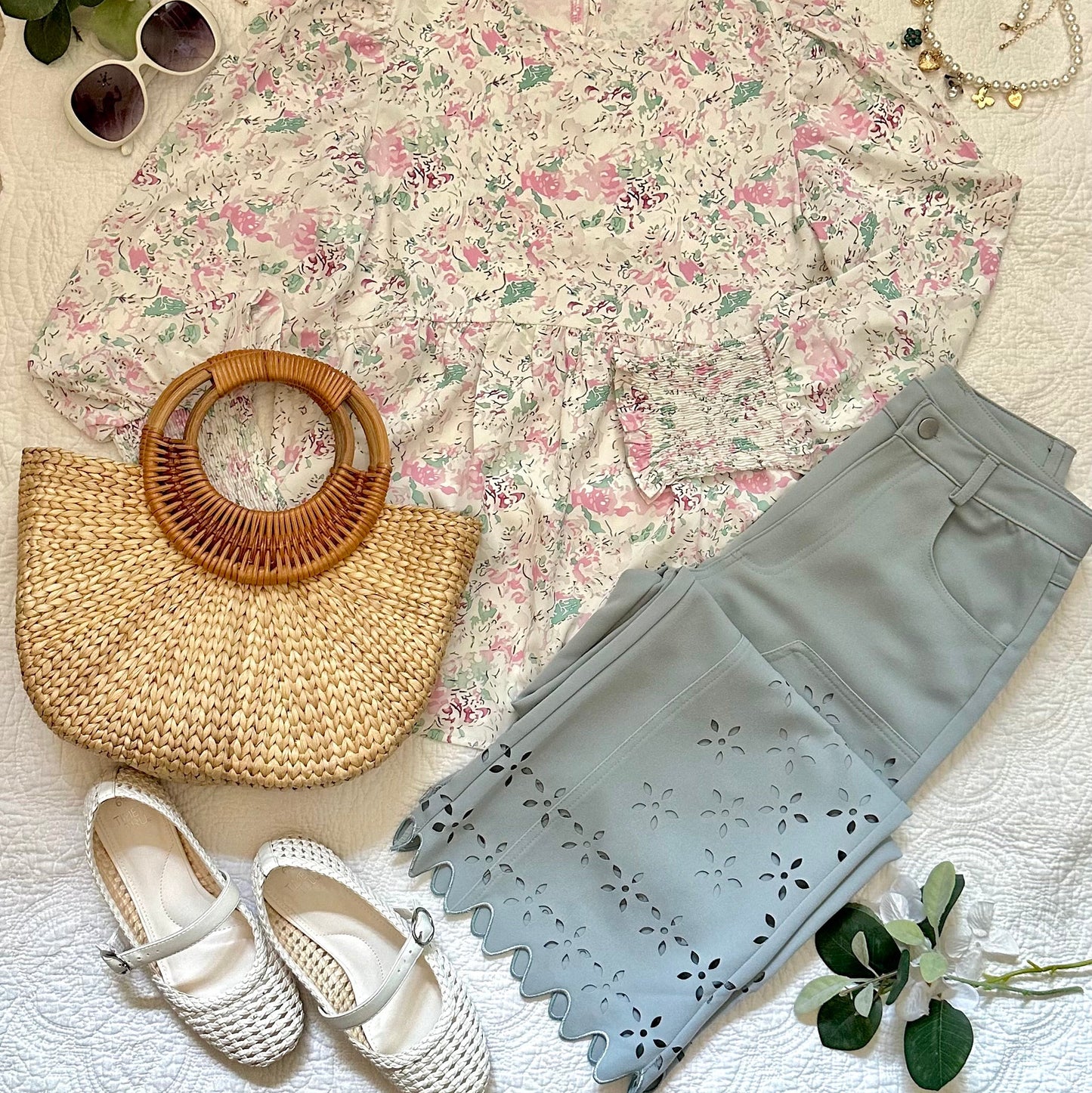 A styled flat-lay outfit on a white textured fabric background, featuring a long-sleeve floral blouse, light blue cropped pants with floral eyelet cutouts, white Mary Jane flats, a woven straw handbag with a round wooden handle, sunglasses, a pearl necklace, and green leaves.