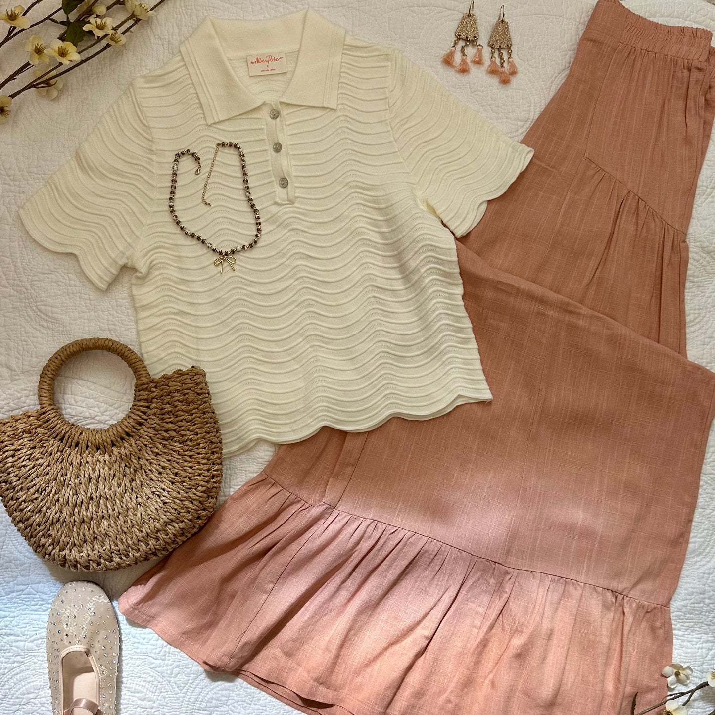 A styled flat-lay outfit featuring a cream ribbed short-sleeve polo shirt with scalloped edges, a dusty rose tiered skirt, a woven straw handbag, beige flats, and dangling earrings arranged on a white textured blanket.