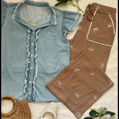 A styled flat-lay of a two-piece clothing set featuring a light blue denim short-sleeve top with ruffled trim and matching brown pants with white embroidered bow details, arranged on a white textured blanket with a woven bag, white shoes, and green leaves.