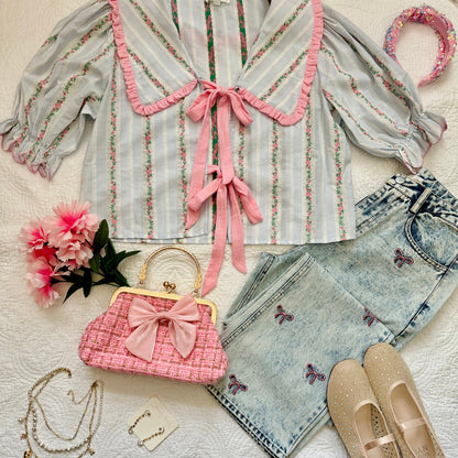 A styled flat-lay outfit on a white textured blanket featuring a light gray and white striped blouse with pink ruffle trim and tie details, light-wash denim shorts with embroidered butterflies, beige slip-on shoes, a pink quilted handbag with a large bow, layered necklaces, earrings, and a pink floral headband.