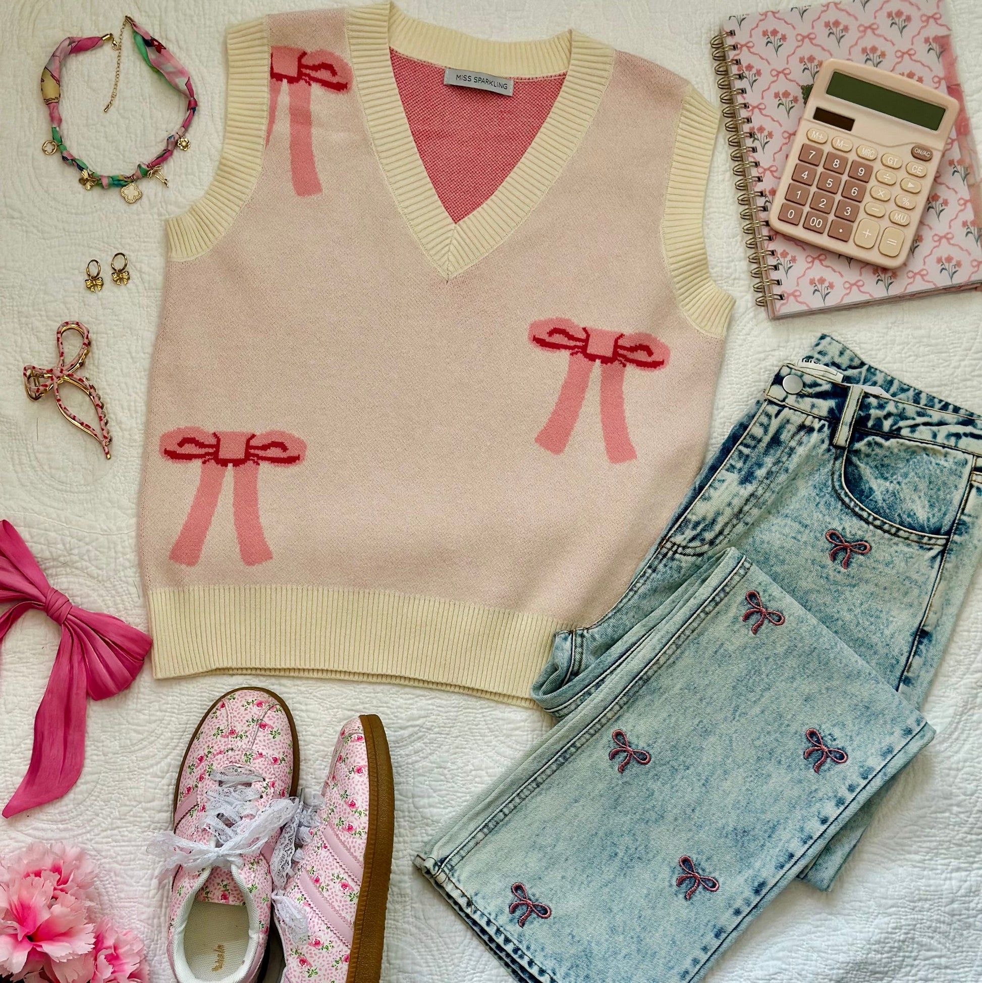 A styled flat-lay outfit on a white textured blanket featuring a cream knit sweater vest with pink bow details, light-wash denim jeans with butterfly embroidery, pink floral sneakers, a pink hair tie, jewelry, a notebook, and a calculator.