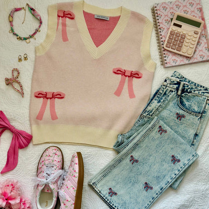A styled flat-lay outfit on a white textured blanket featuring a cream knit sweater vest with pink bow details, light-wash denim jeans with butterfly embroidery, pink floral sneakers, a pink hair tie, jewelry, a notebook, and a calculator.