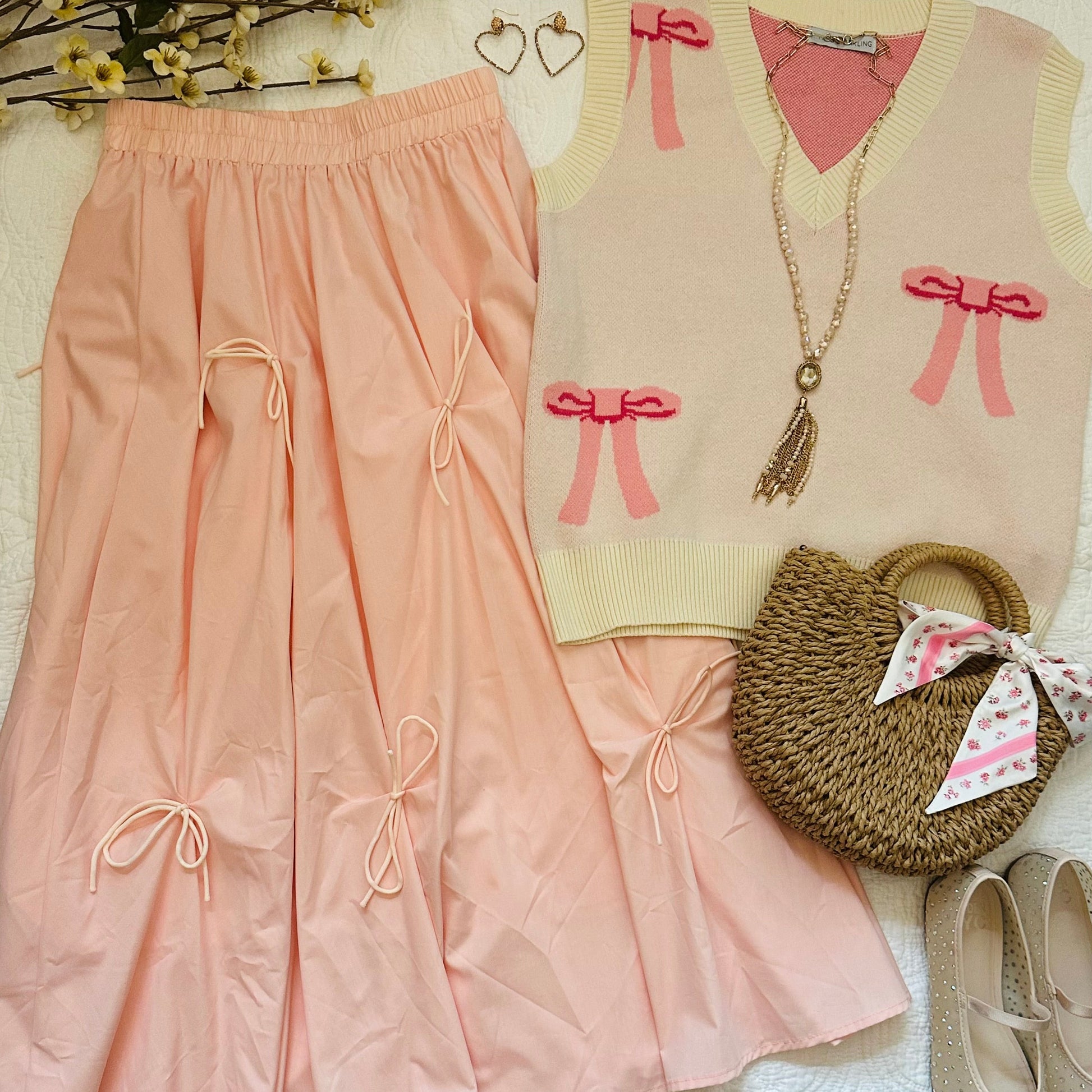 A styled flat-lay outfit featuring a two-piece set of light peach-pink pleated pants and a matching sleeveless knit vest with pink bow details, styled with a woven handbag, gold jewelry, beige shoes, and decorative flowers on a white textured background.