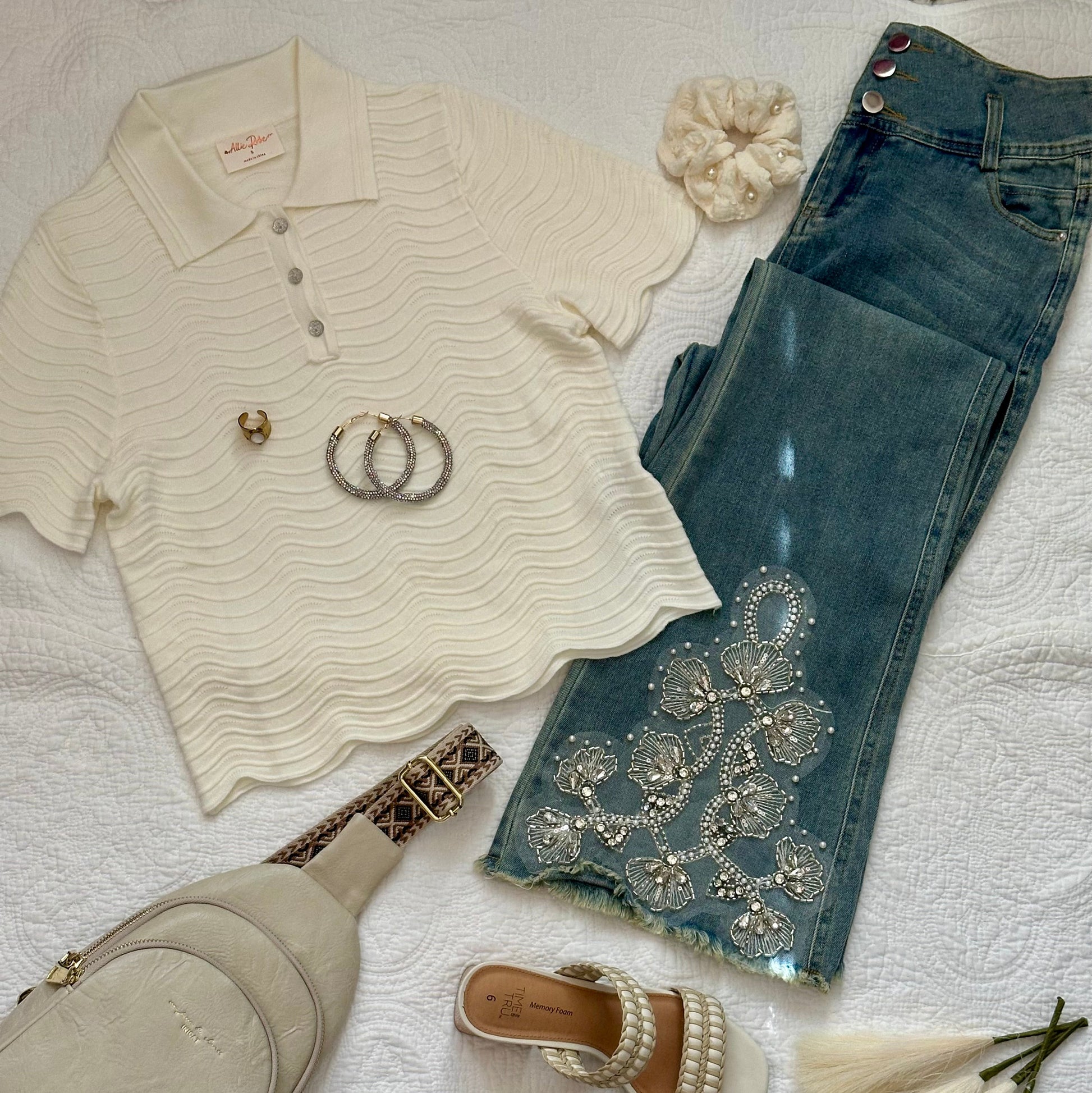 A styled flat-lay outfit on a white textured fabric background, featuring a cream ribbed short-sleeve polo shirt, blue jeans with silver rhinestone embellishments, a light beige crossbody bag, pearl-strap heeled sandals, and a cream scrunchie.