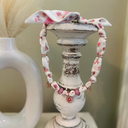 A fabric beaded necklace with pink floral patterns and small pink beads, draped over a distressed white candle holder.