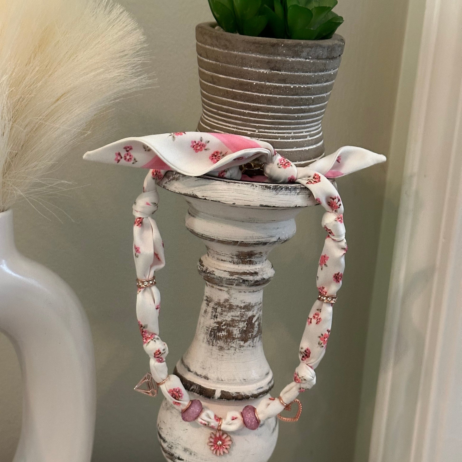 A close-up of a white distressed decorative candle holder with a floral fabric charm necklace holding a small potted green plant, against an indoor background.