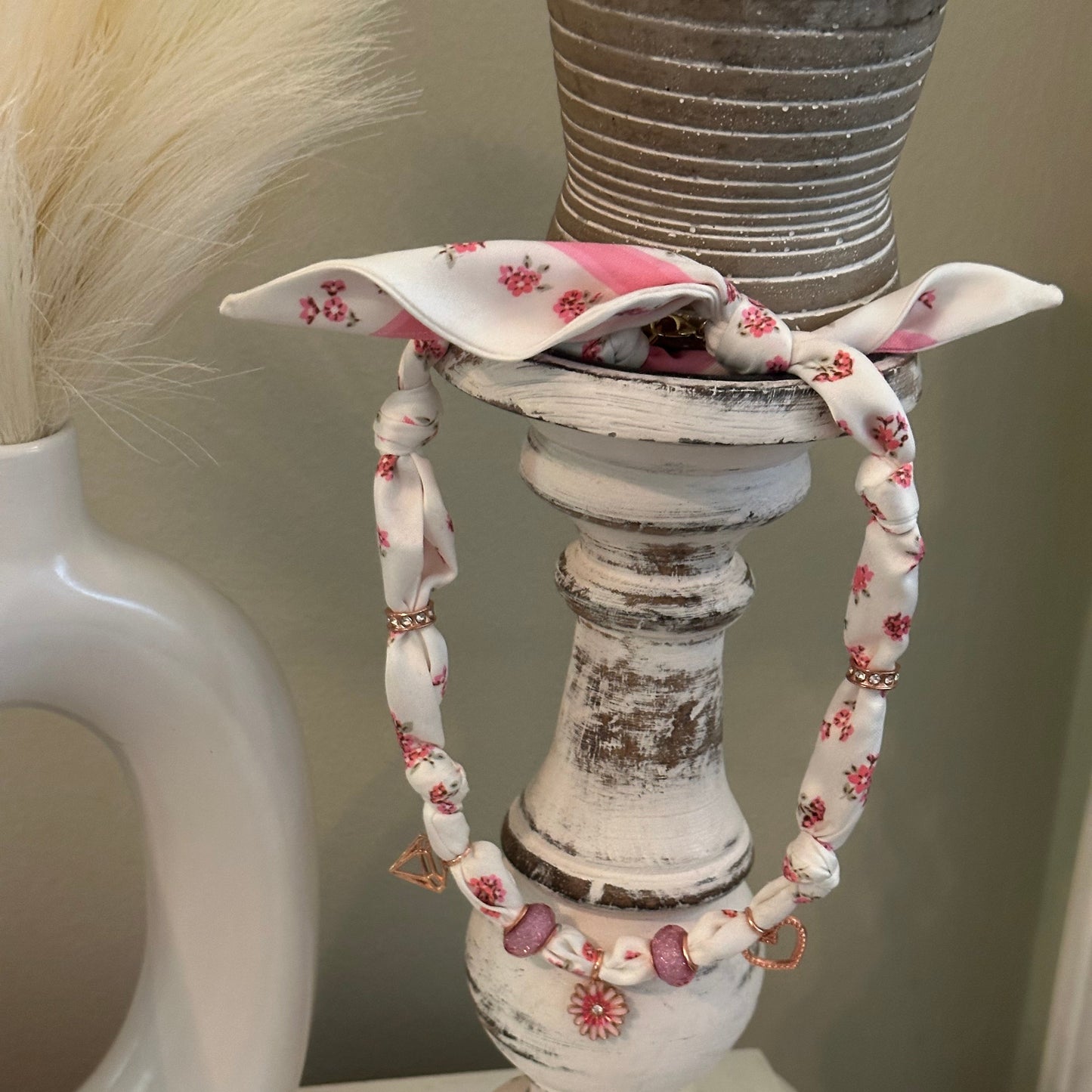 A close-up of a distressed white decorative candle holder or lamp post with a floral fabric charm necklace draped around it. The fabric is white with small pink and red flowers and has a pink bow at the top.