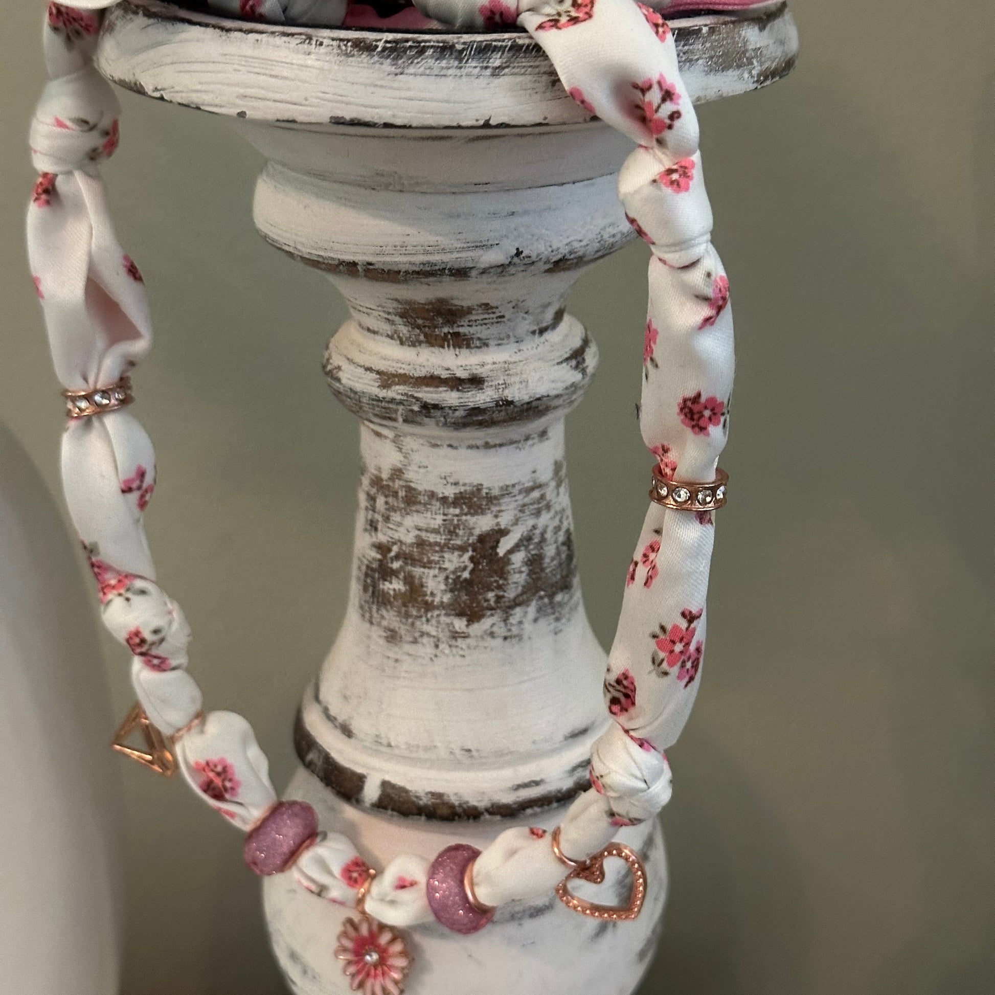 A close-up of a white fabric charm necklace with a pink and white floral print, styled on a distressed white wooden  candlestick.