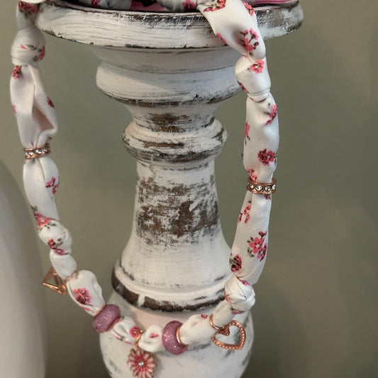 A close-up of a white fabric charm necklace with a pink and white floral print, styled on a distressed white wooden  candlestick.