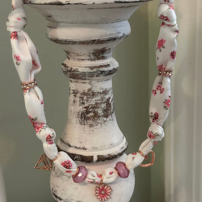A close-up of a handmade fabric and bead necklace draped over a distressed white wooden candlestick holder. The necklace features knotted white fabric segments with a pink floral pattern, accented by pink and purple beads, rose gold-toned metal rings, and a small rose gold geometric charm.