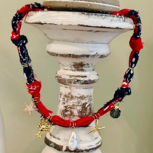 A handmade bohemian-style necklace made from red and navy blue patterned fabric bands, decorated with small gold starfish, shell charms, and a teardrop pearl pendant, displayed on a distressed white wooden stand.