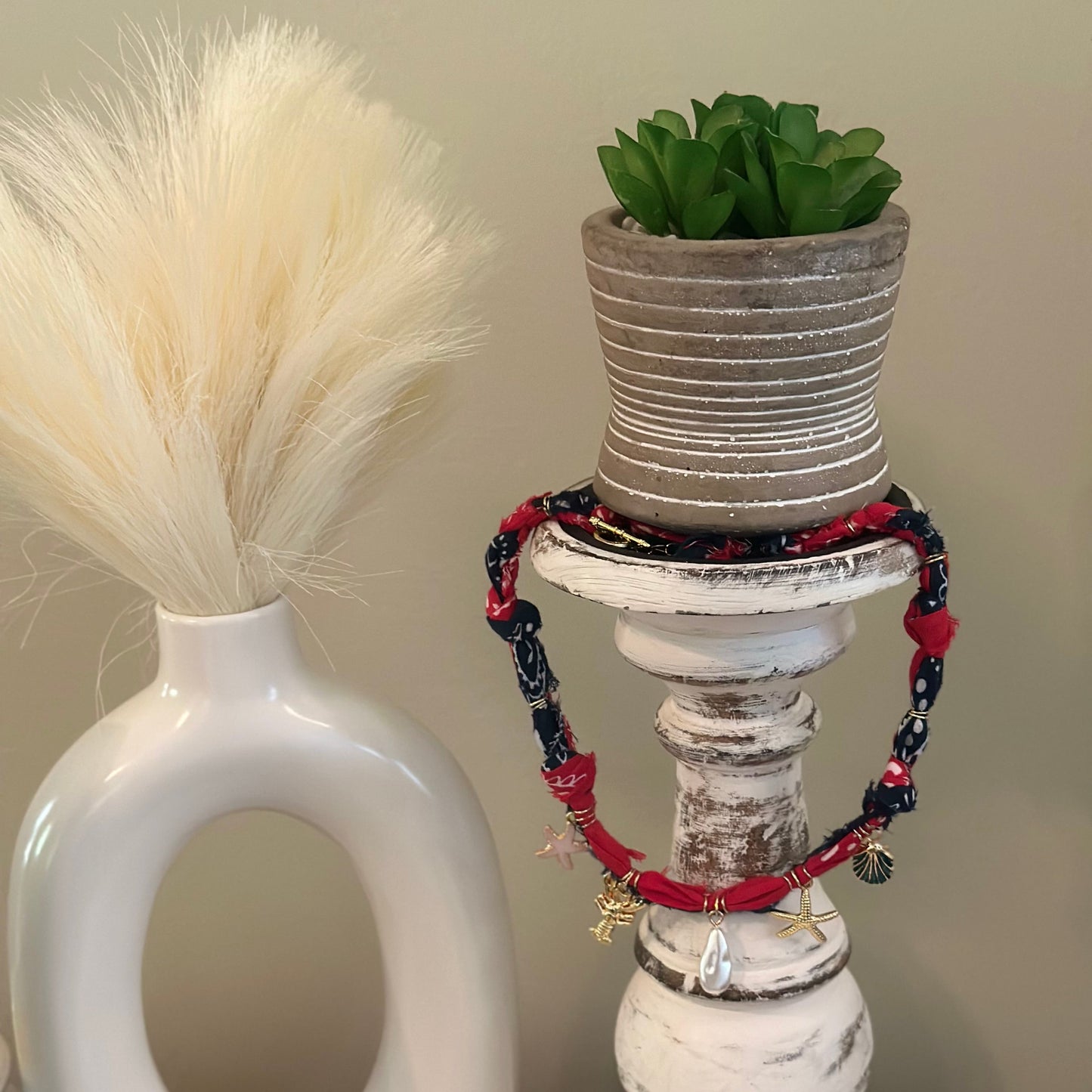 A set of red and navy blue beaded bracelets with small gold charms, draped around a distressed white candlestick holder with a small potted succulent on top.