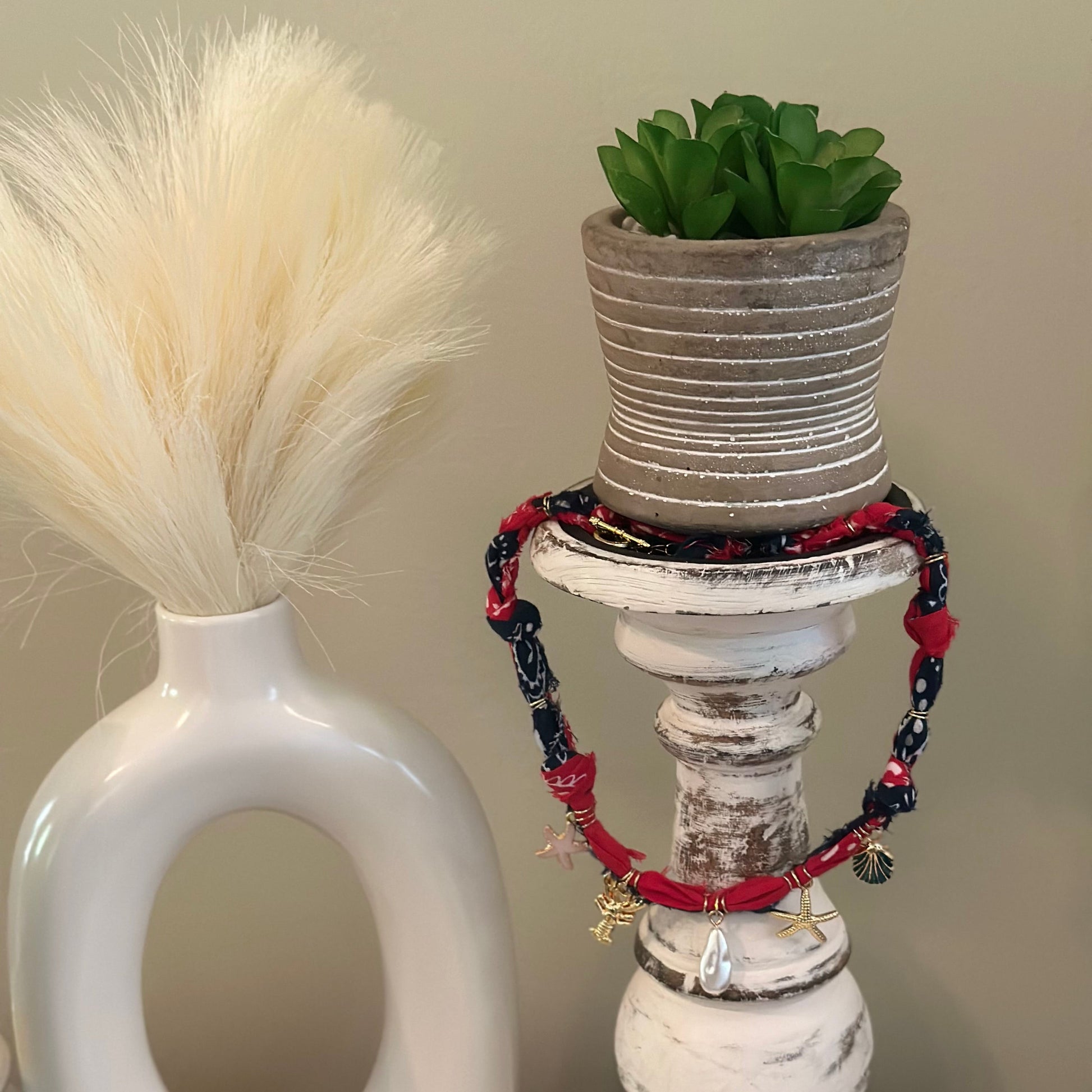 A set of red and navy blue beaded bracelets with small gold charms, draped around a distressed white candlestick holder with a small potted succulent on top.