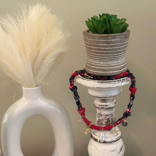 A set of red and navy blue beaded bracelets with small gold charms, draped around a distressed white candlestick holder with a small potted succulent on top.