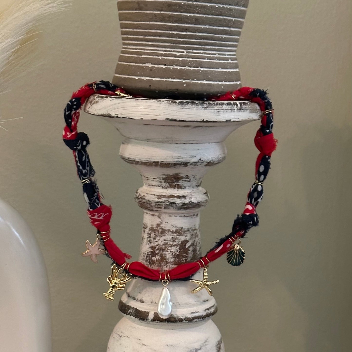 A red and navy blue beaded charm necklace with gold-toned pendants, displayed on a distressed white wooden candlestick holder with a small potted succulent on top.