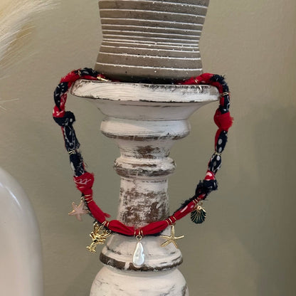 A red and navy blue beaded charm necklace with gold-toned pendants, displayed on a distressed white wooden candlestick holder with a small potted succulent on top.