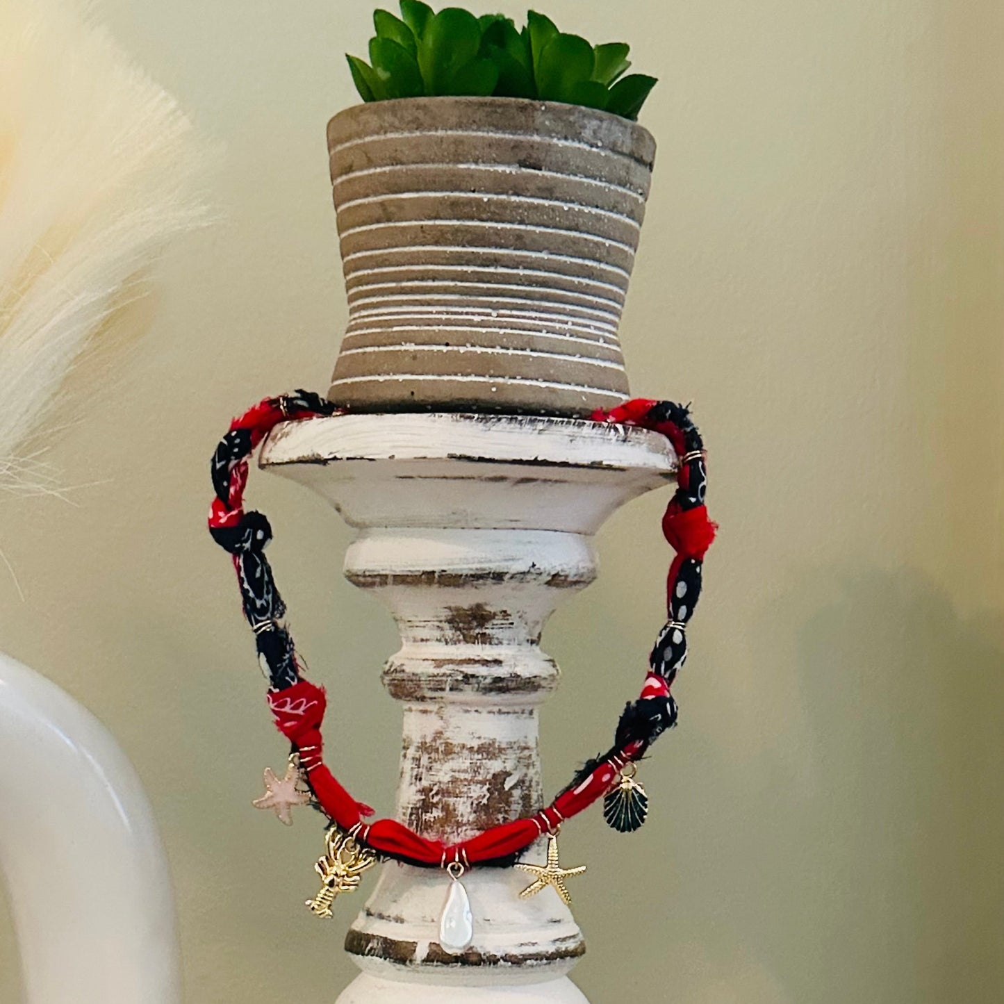A red and black beaded bracelet with starfish and pearl charms draped over a distressed white wooden candle holder, with a small potted succulent on top.