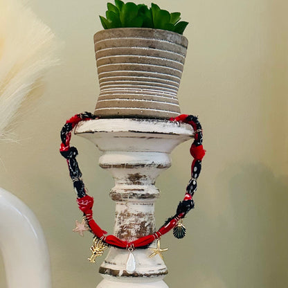 A red and black beaded bracelet with starfish and pearl charms draped over a distressed white wooden candle holder, with a small potted succulent on top.