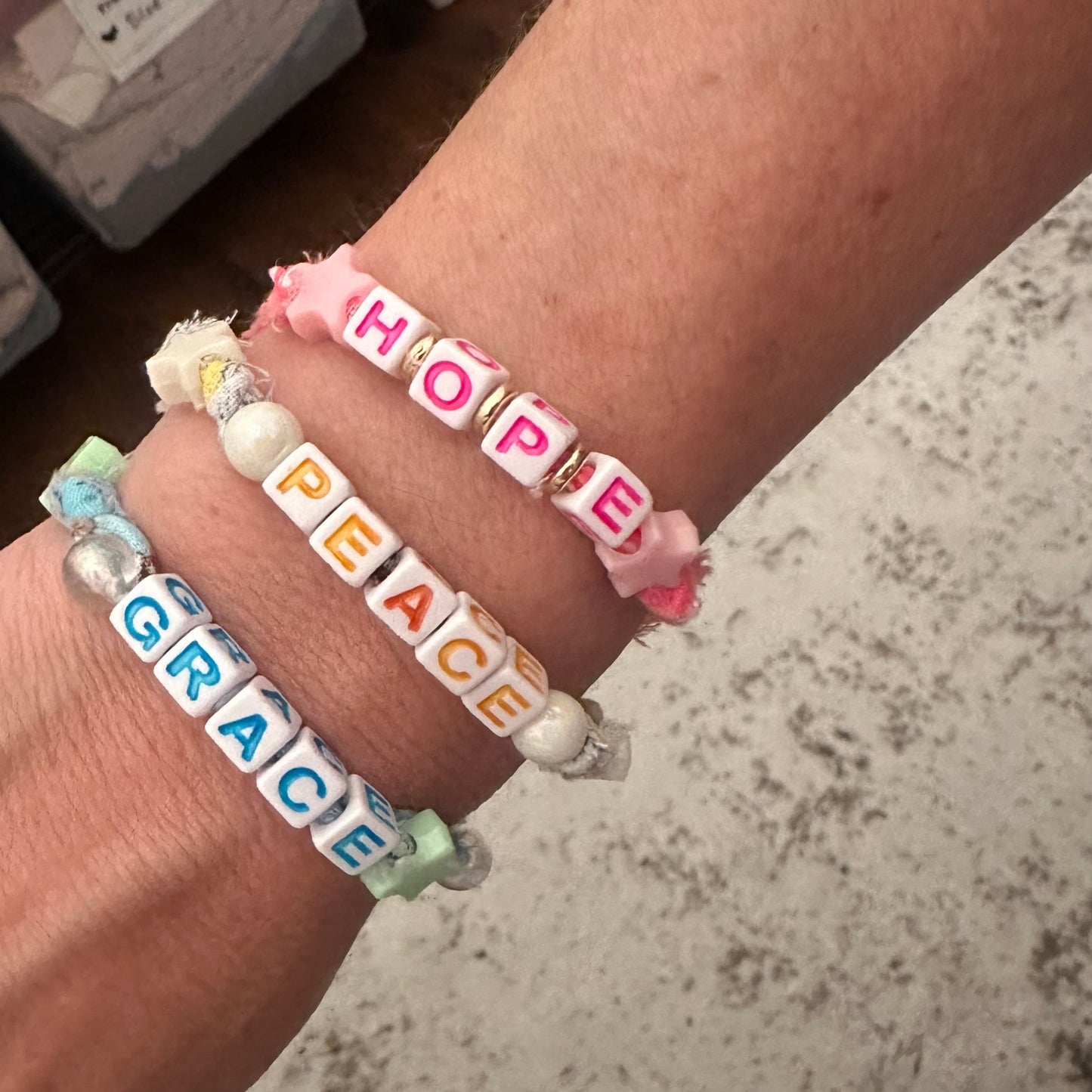 Three beaded letter bracelets on a person's wrist, spelling out HOPE, PEACE, and GRACE, with pastel pink, white, and light blue cords and colorful letter beads.
