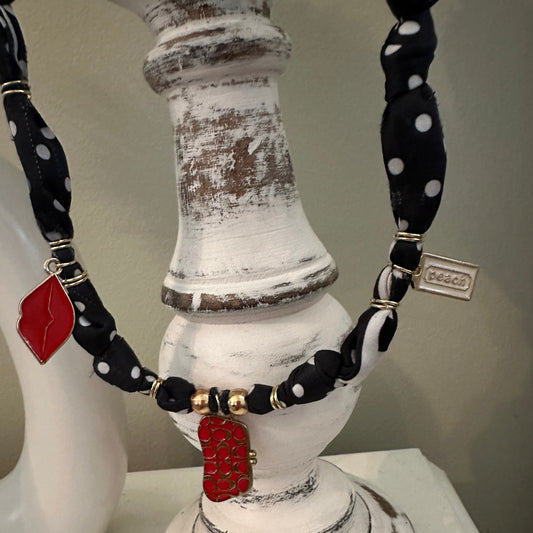 A close-up of a necklace displayed on a distressed white decorative stand. The necklace features black fabric with white polka dots, black beads, gold-toned spacer beads, and two red enamel lip-shaped charms.
