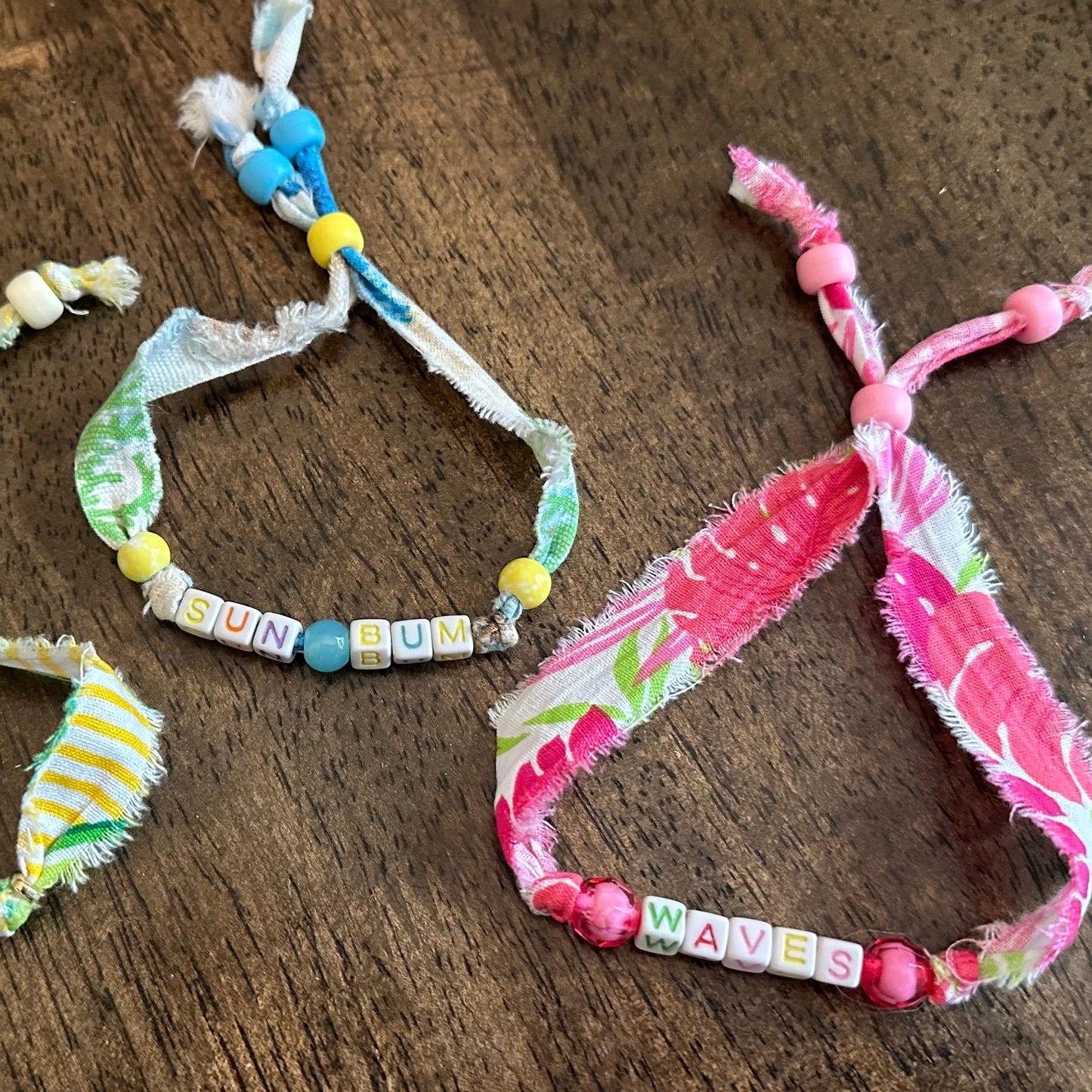 Several handmade fabric charm bracelets with colorful beads and frayed ties are arranged on a dark wooden surface.