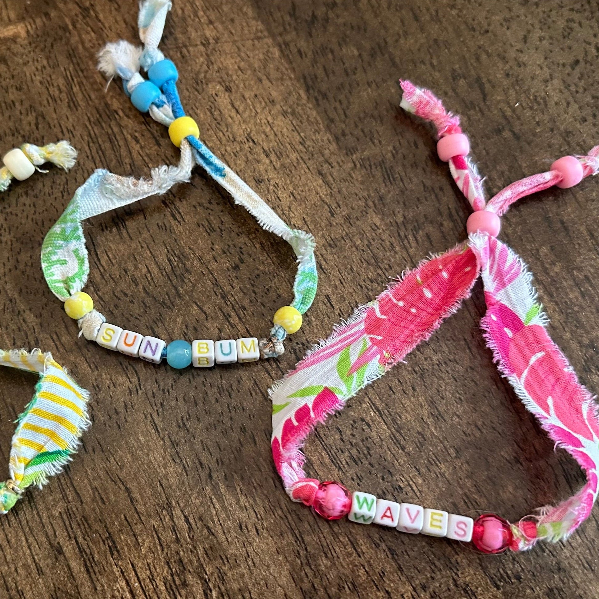 Several handmade fabric charm bracelets with colorful beads and frayed ties are arranged on a dark wooden surface.