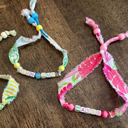 Several handmade fabric charm bracelets with colorful beads and frayed ties are arranged on a dark wooden surface.
