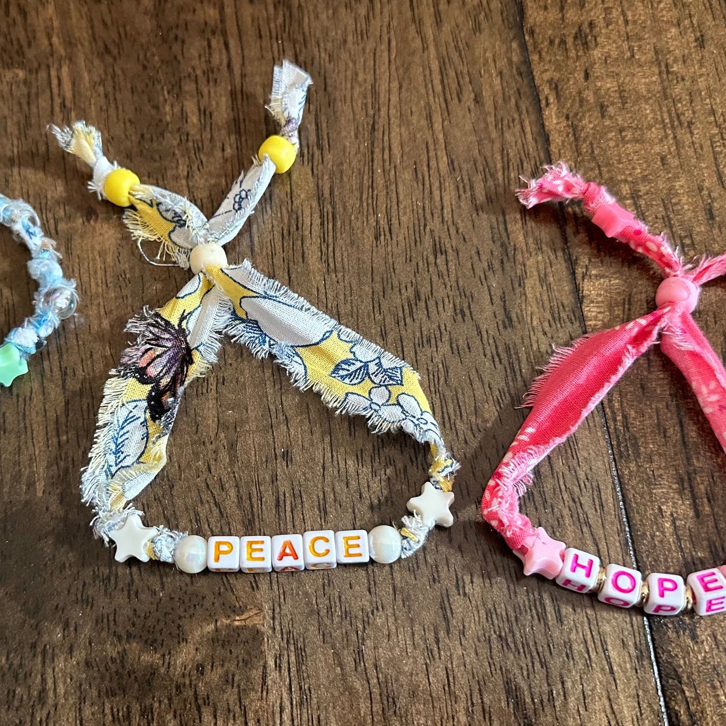 Three handmade fabric friendship bracelets with letter beads spelling out GRACE, PEACE, and HOPE, laid out on a dark wooden surface.