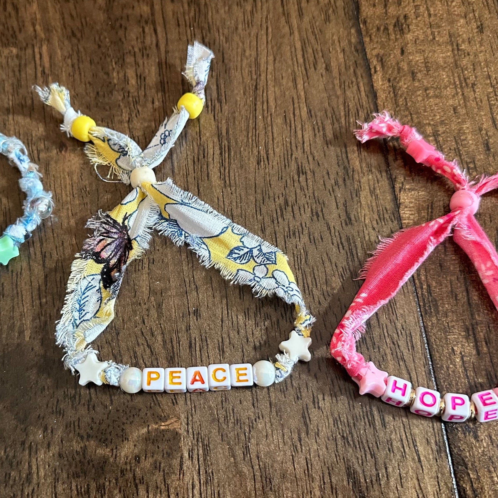 Three handmade fabric friendship bracelets with letter beads spelling out GRACE, PEACE, and HOPE, laid out on a dark wooden surface.