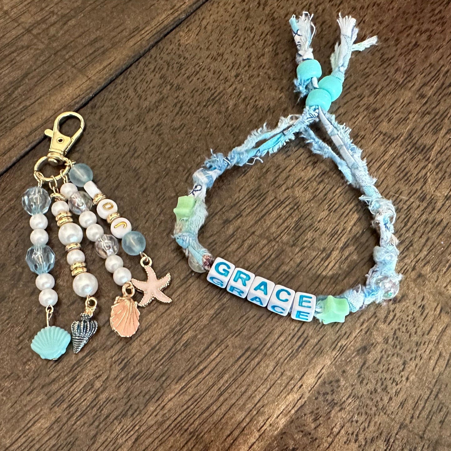 A handmade beaded keychain and matching bracelet on a dark wood background. The keychain features white and light blue beads, a gold clasp, and small charms including a starfish and a seashell.