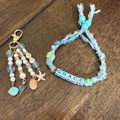 A handmade beaded keychain and matching bracelet on a dark wood background. The keychain features white and light blue beads, a gold clasp, and small charms including a starfish and a seashell.