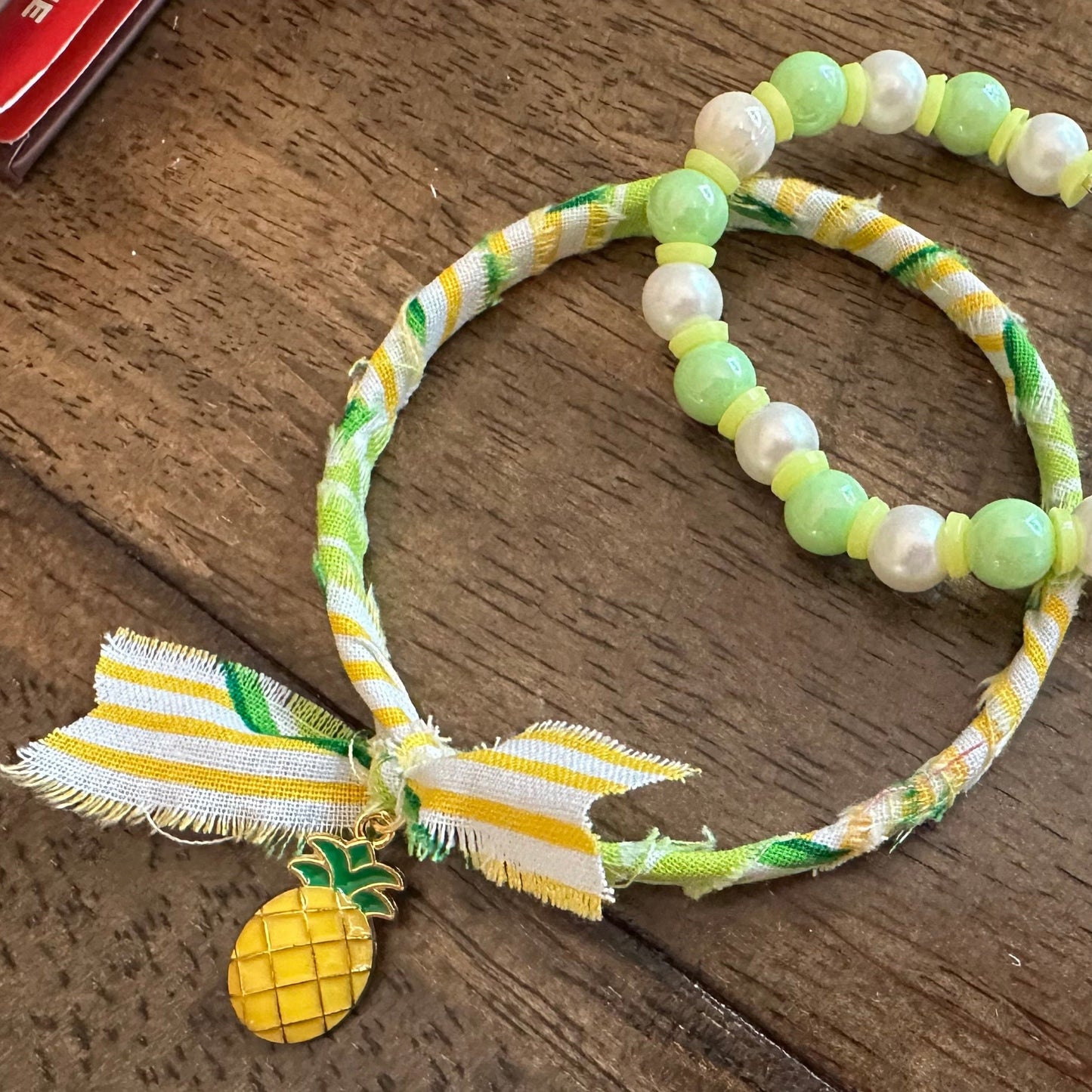 A beaded bracelet with light green and white pearls, a yellow and white striped ribbon bow, and a small pineapple charm, displayed on a dark wooden surface.