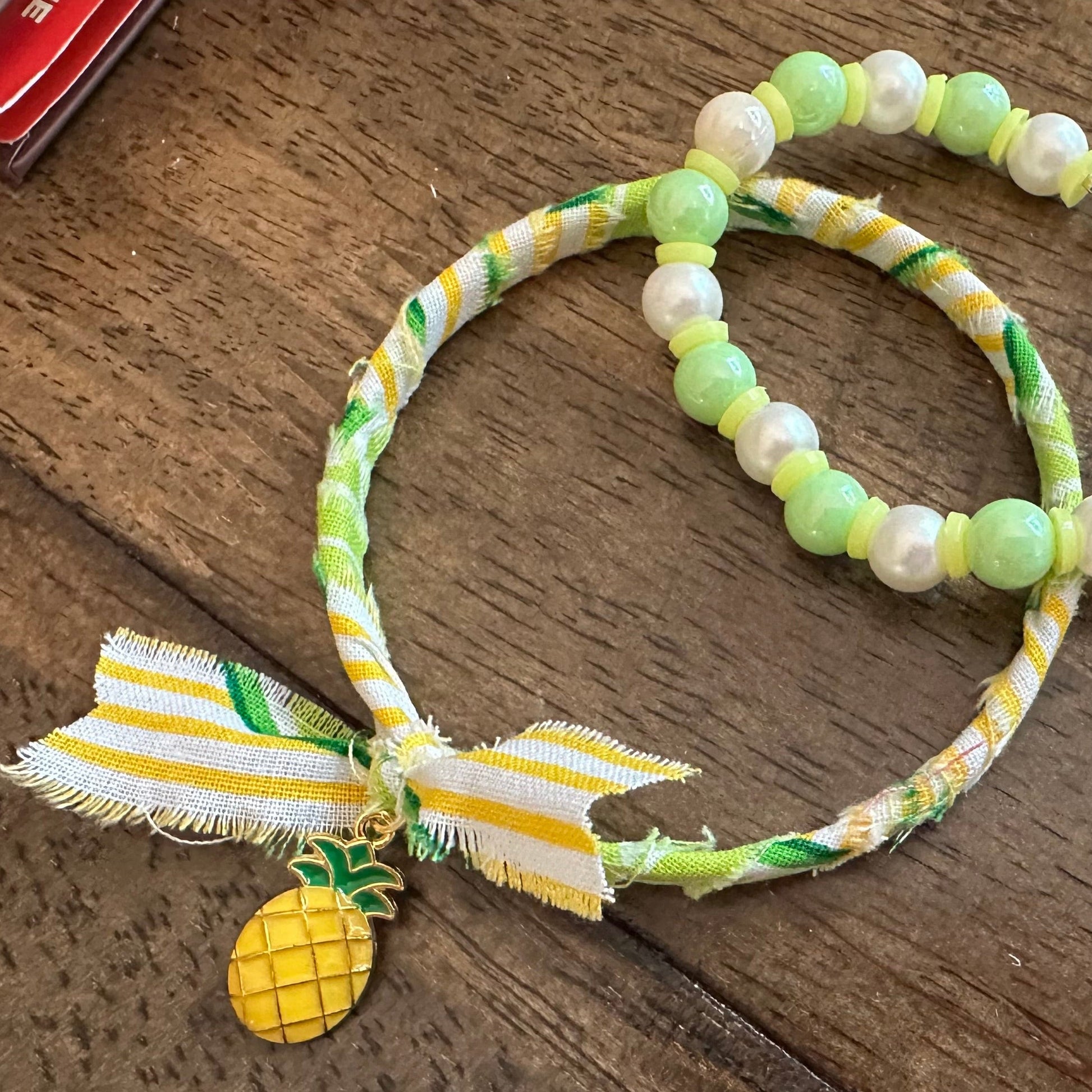 A beaded bracelet with light green and white pearls, a yellow and white striped ribbon bow, and a small pineapple charm, displayed on a dark wooden surface.