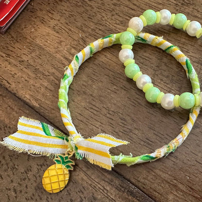 A beaded bracelet with light green and white pearls, a yellow and white striped ribbon bow, and a small pineapple charm, displayed on a dark wooden surface.