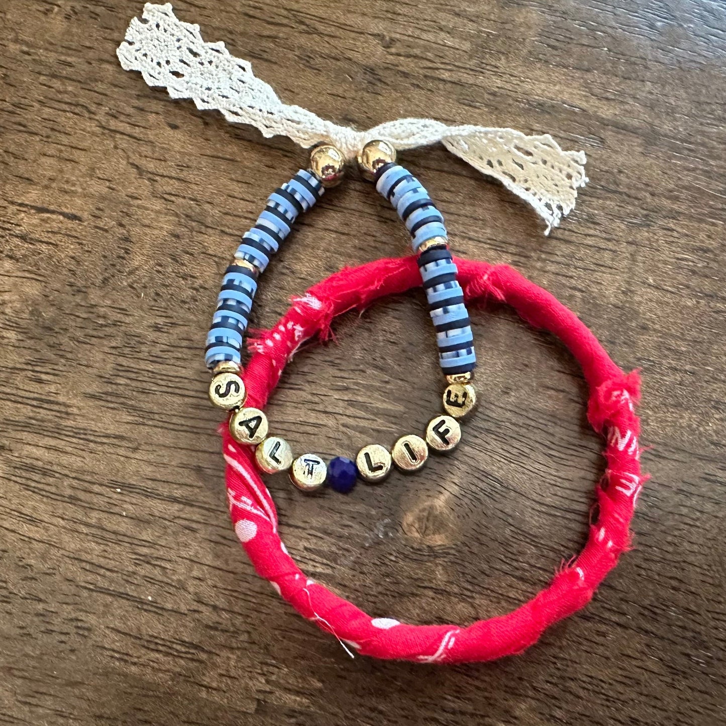 Three handmade beaded bracelets displayed on a dark wooden table. One bracelet is made of bright red fabric-wrapped beads with a white lace tassel.