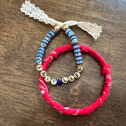 Three handmade beaded bracelets displayed on a dark wooden table. One bracelet is made of bright red fabric-wrapped beads with a white lace tassel.