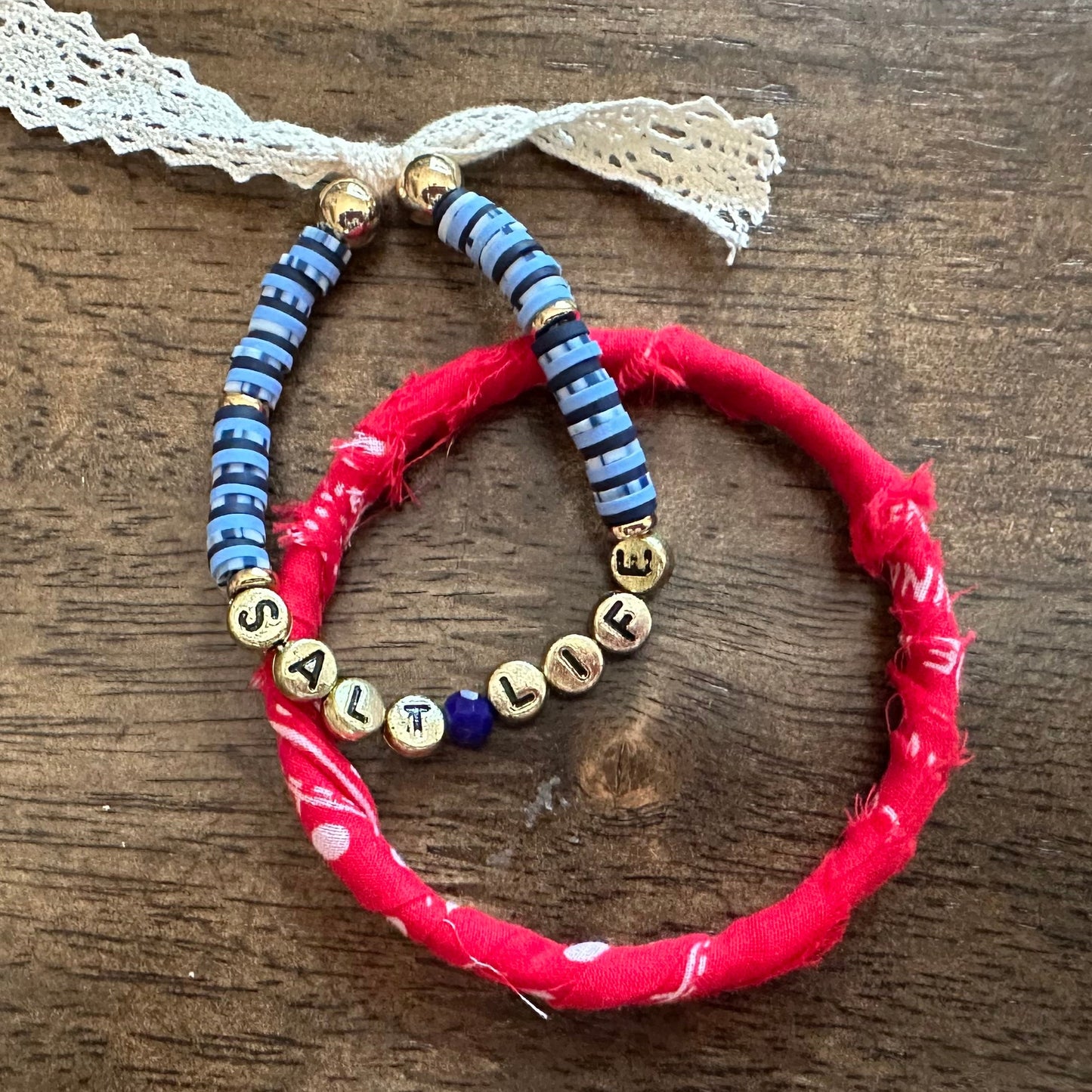 Two handmade beaded bracelets on a dark wood background. One bracelet is bright red with a small white bead accent, and the other is blue with gold letter beads spelling 'LIFE' and blue spacer beads.