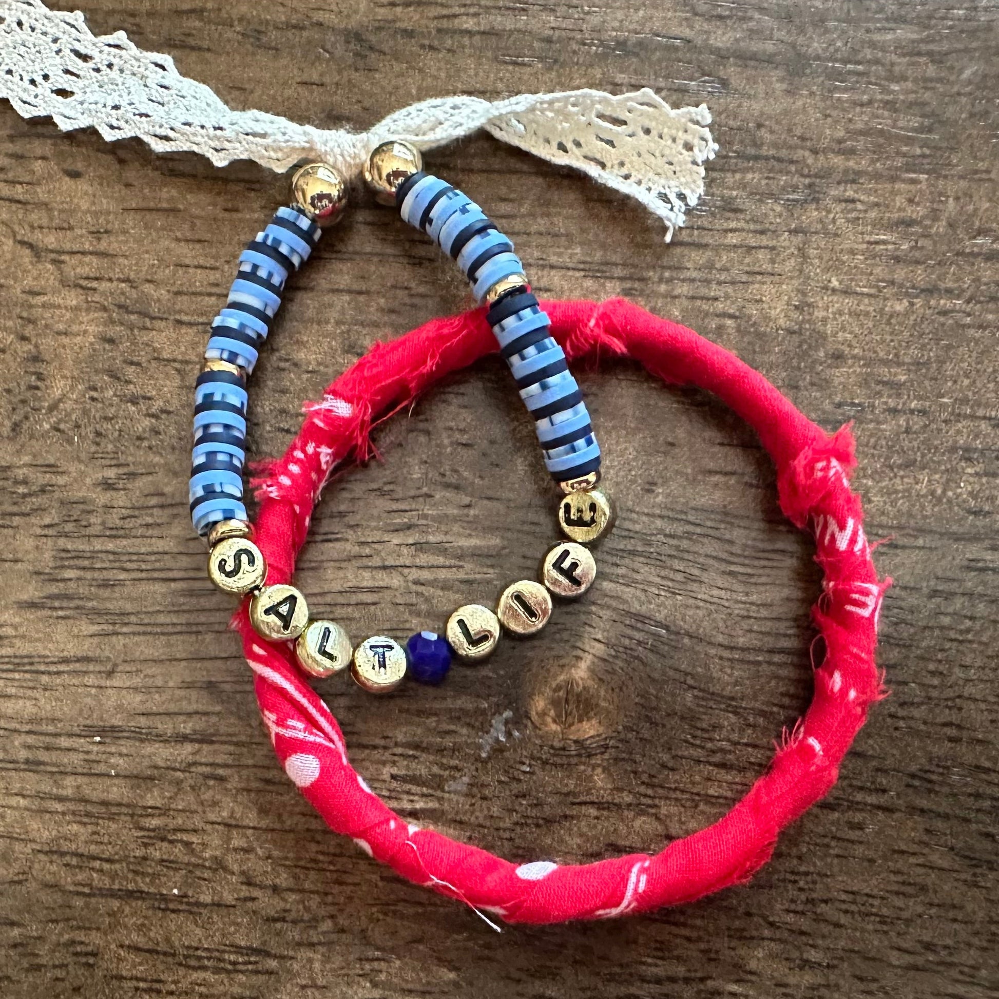 Two handmade beaded bracelets on a dark wood background. One bracelet is bright red with a small white bead accent, and the other is blue with gold letter beads spelling 'LIFE' and blue spacer beads.